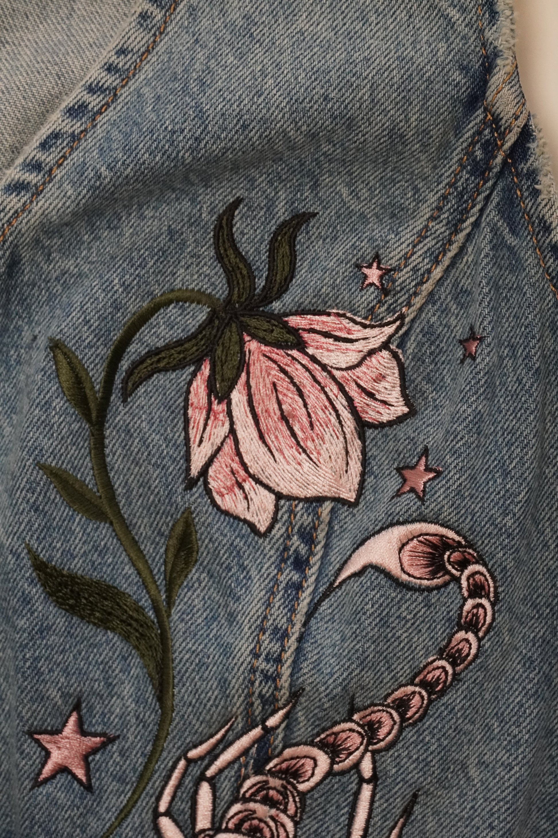 Close-up of a blue denim garment featuring embroidered details, including a pink flower with green leaves, pink stars, and a chain motif.
