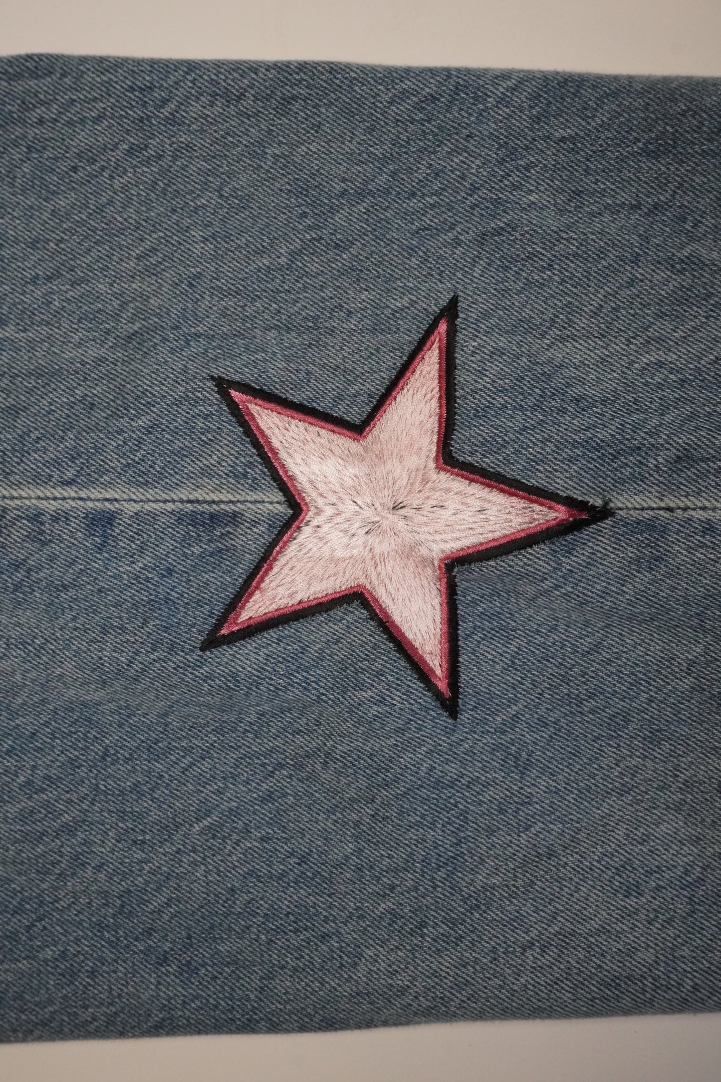 A close-up of blue denim fabric with a five-pointed star patch in the center. The star has a light pink center and a dark red border, outlined with black stitching.