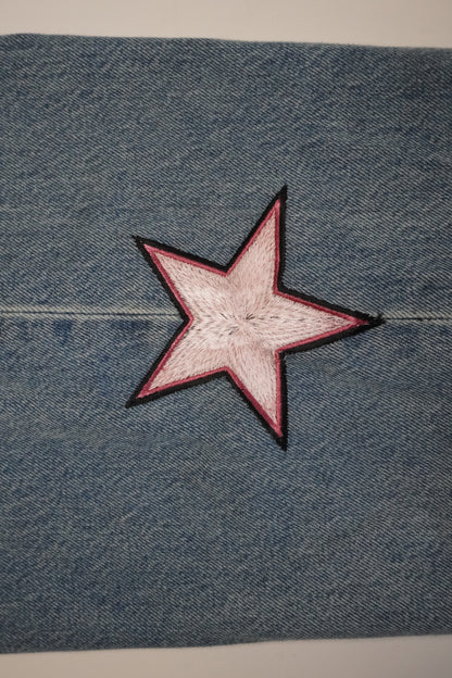 A close-up of blue denim fabric with a five-pointed star patch in the center. The star has a light pink center and a dark red border, outlined with black stitching.