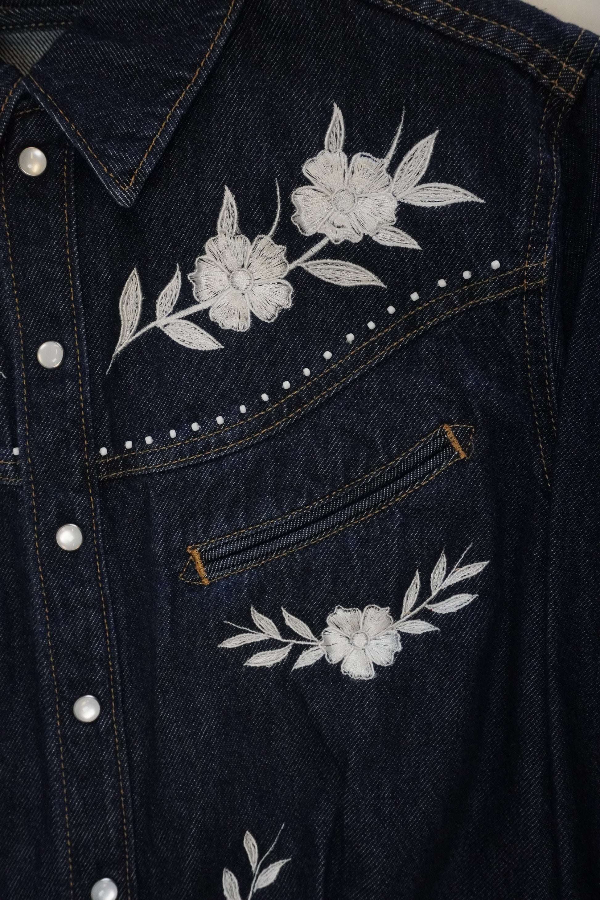 Close-up of dark blue denim jeans showing the back pocket with white floral embroidery, silver metal rivets, and contrast stitching.