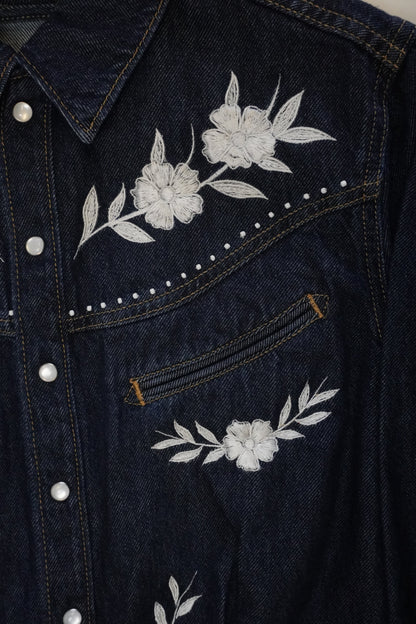 Close-up of dark blue denim jeans showing the back pocket with white floral embroidery, silver metal rivets, and contrast stitching.