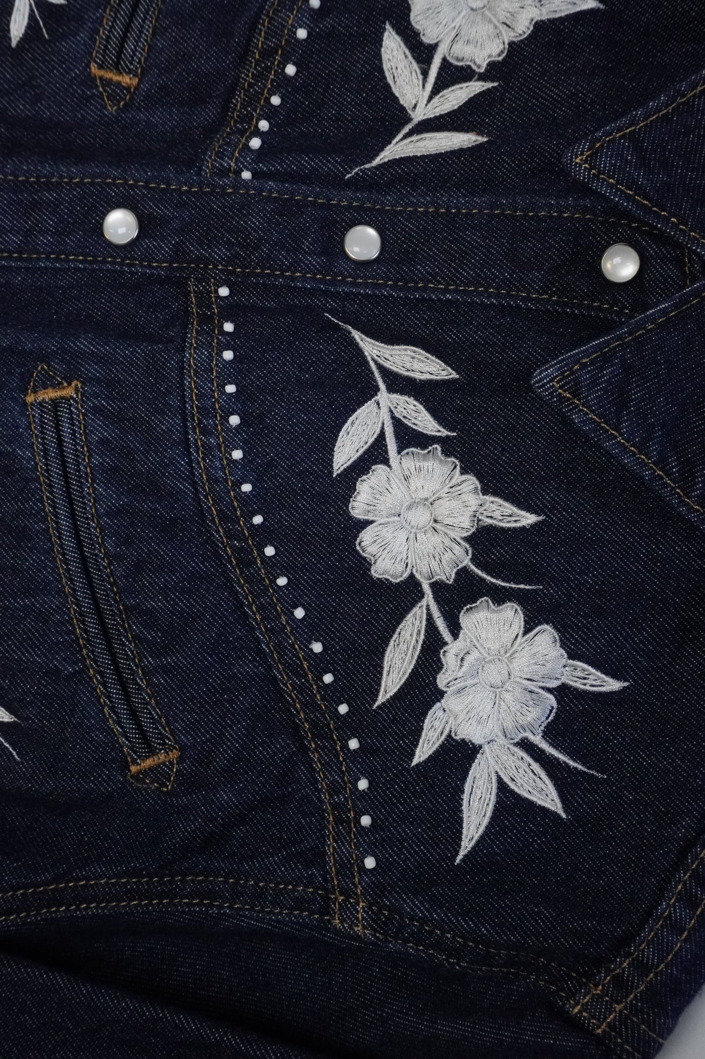Close-up of a dark indigo denim jacket featuring white embroidered flowers and leaves on the front, accented with white rhinestone trim and silver snap buttons.