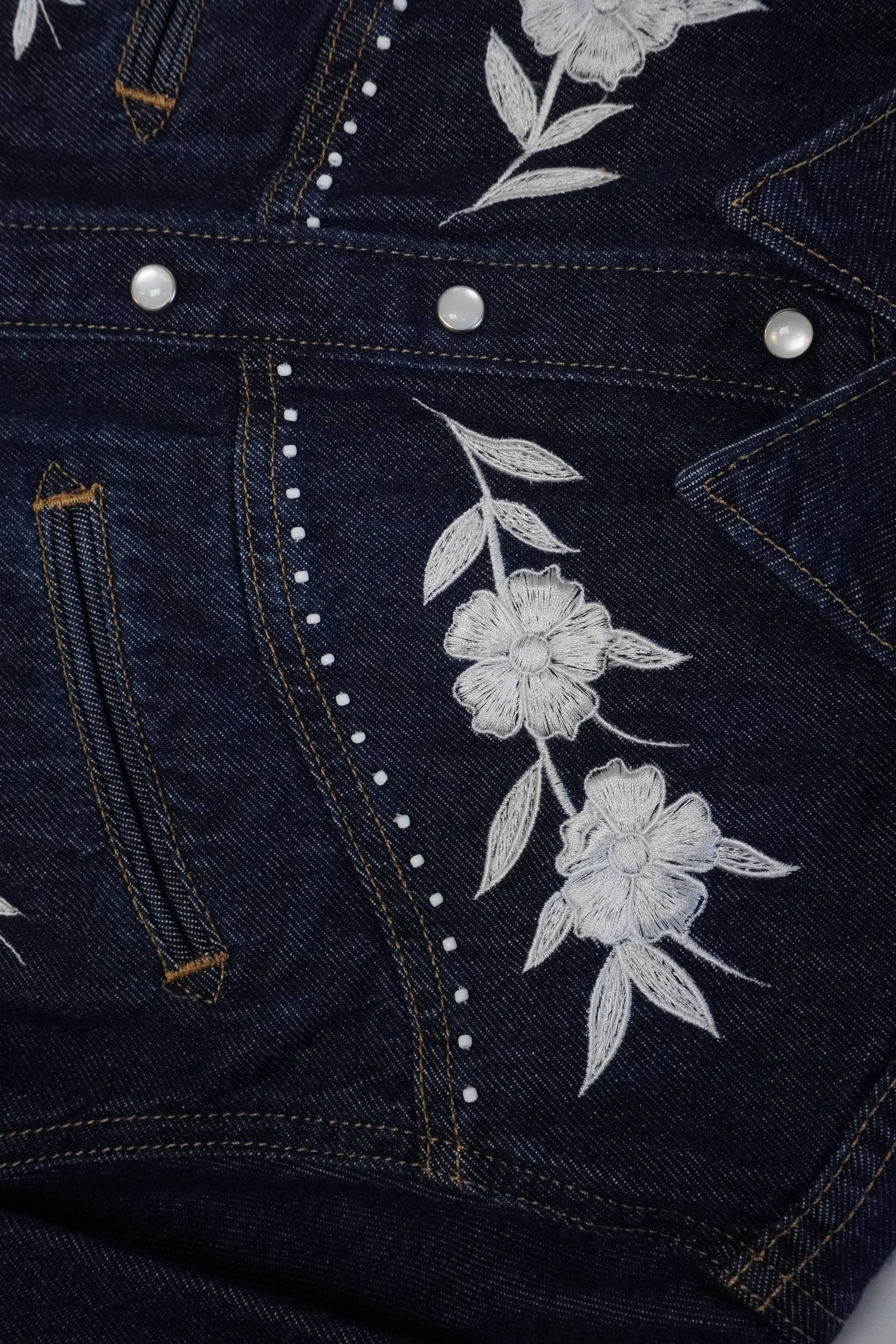 Close-up of a dark indigo denim jacket featuring white embroidered flowers and leaves on the front, accented with white rhinestone trim and silver snap buttons.
