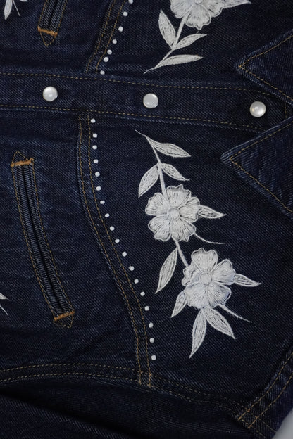 Close-up of a dark indigo denim jacket featuring white embroidered flowers and leaves on the front, accented with white rhinestone trim and silver snap buttons.