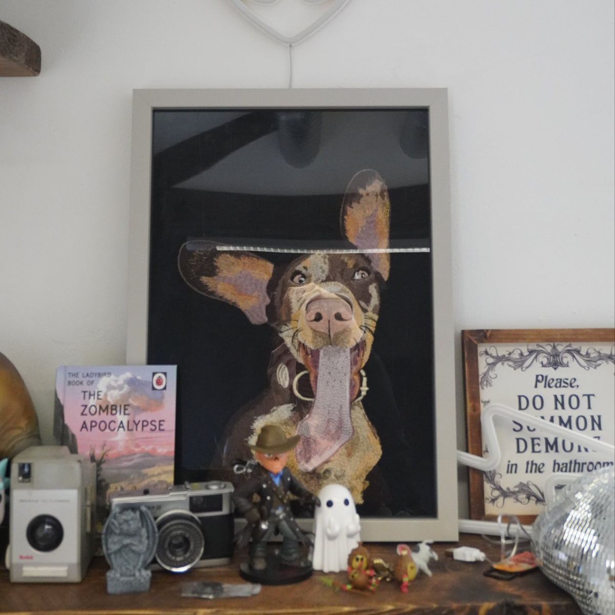 A framed art print featuring a painted dog with its tongue sticking out, displayed on a wooden shelf alongside a camera, a small ghost figurine, a book, and other decorative items against a white wall.