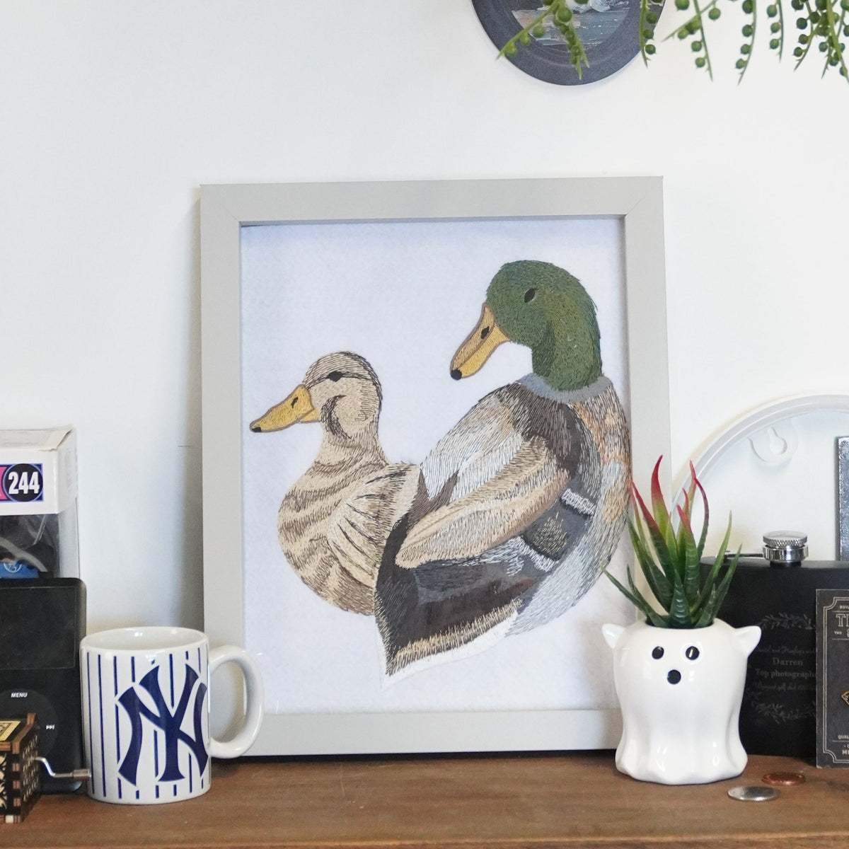 A framed watercolor painting of two ducks displayed on a wooden shelf in a styled home interior, surrounded by plants, a New York Yankees mug, and other decor items.
