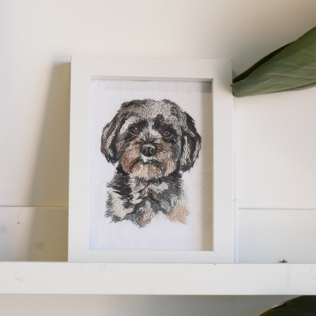 A framed watercolor portrait of a small fluffy dog, displayed on a white shelf against a white wall with a green leaf in the background.