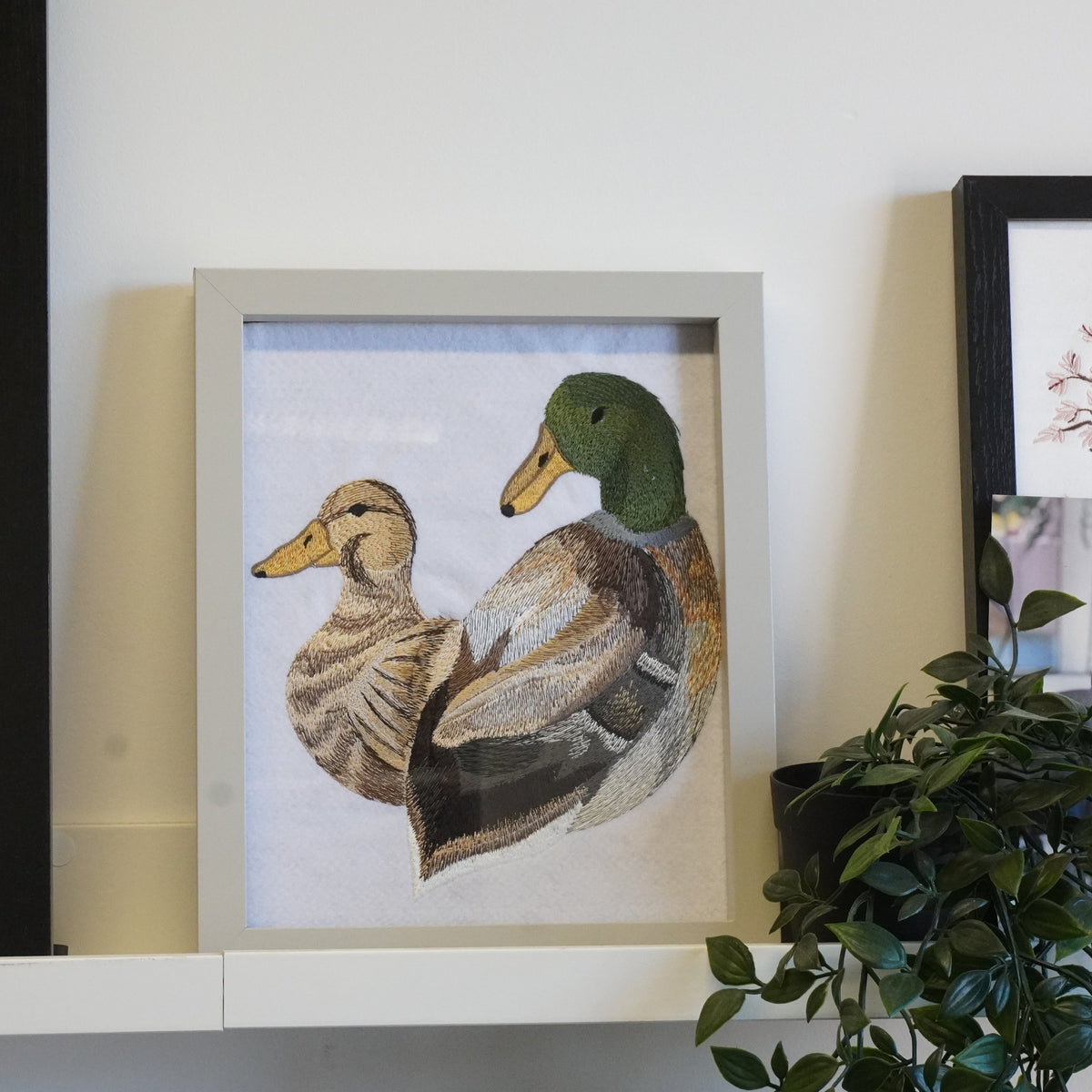 A framed art print featuring a mother duck and her duckling, styled on a white shelf next to a potted green plant and other framed prints.