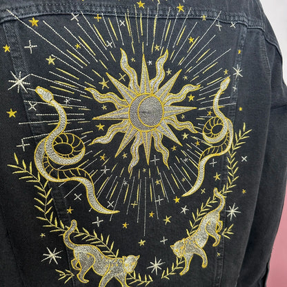 Black denim jacket with gold embroidered design of a sun, snakes, and cats on a plain background