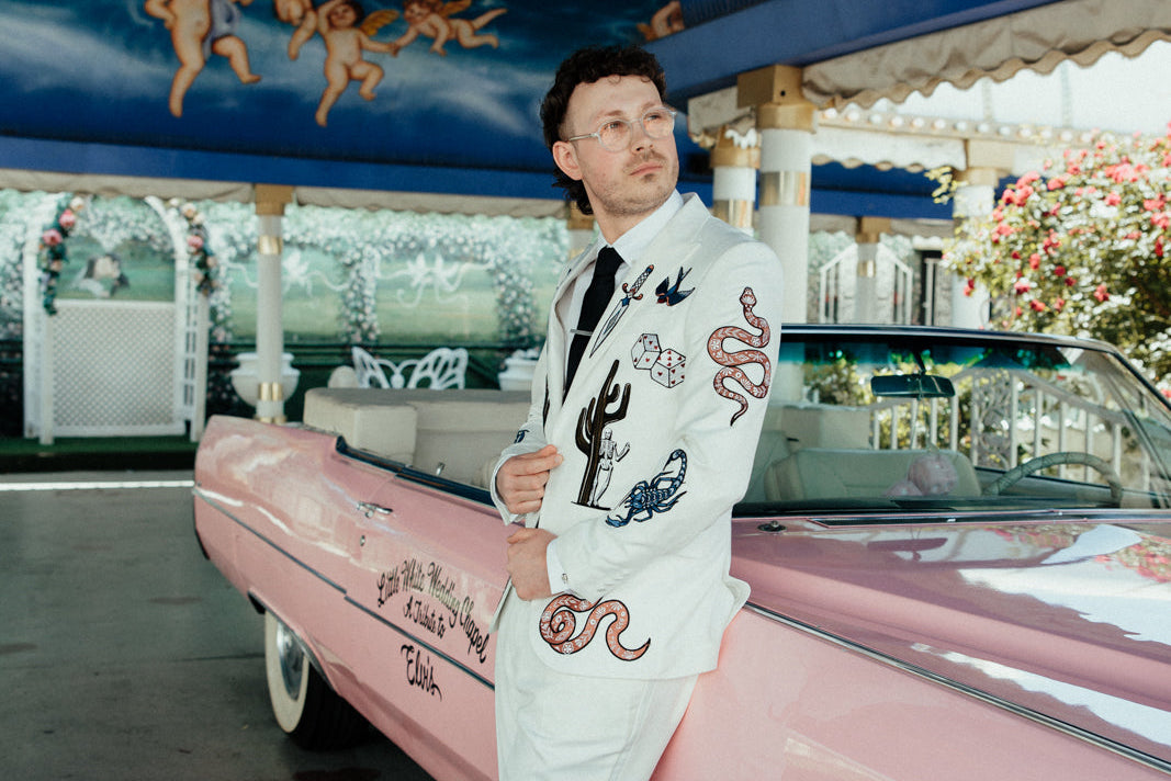 Man in a white suit with colorful embroidery standing next to a pink vintage car under a decorative ceiling.