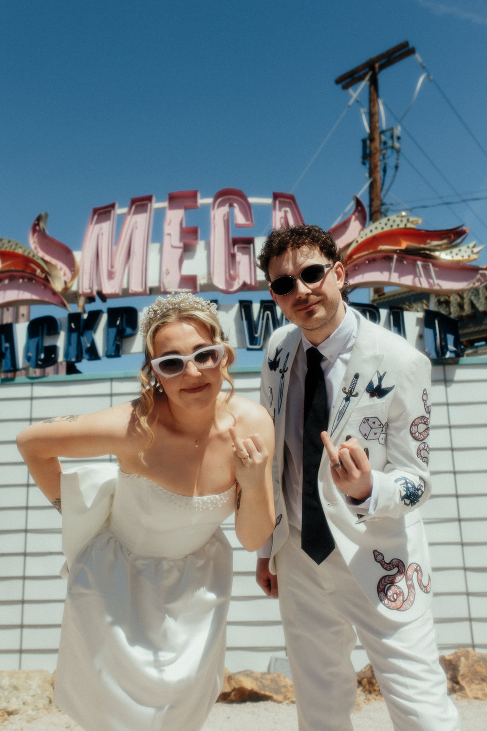 Bride and groom in Vegas neon graveyard museum. The groom is wearing a custom bespoke embroidered white suit with casino and western imagery all over it. 
