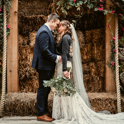 Couple embracing under floral arc and rustic hay bail backdrop. The bride wears a black denim embroidered jean jacket with florals and mountain.