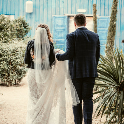 A bride walking away next to her groom, wearing a black denim jacket embroidered on the back with the mountain floral design. Stitched by Beth Wilson Embroidery 