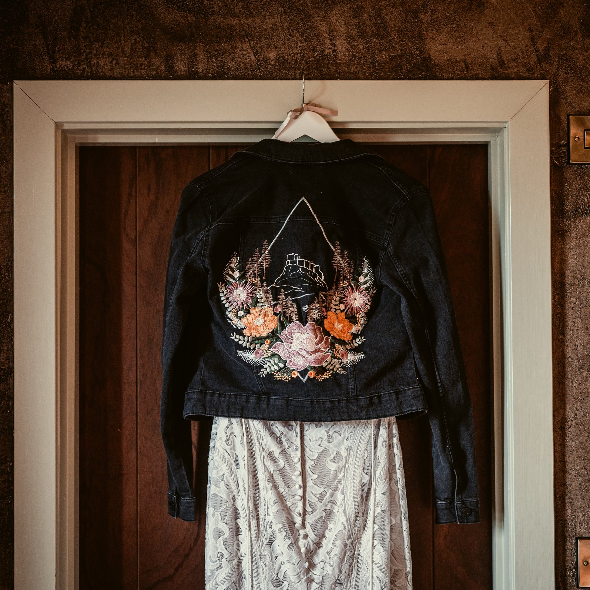 A black denim jacket with a large colorful floral and feather back print, hanging on a white hanger in front of a dark wood door. A white lace skirt is partially visible underneath.