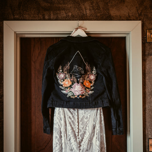 A black denim jacket with a large colorful floral and feather back print, hanging on a white hanger in front of a dark wood door. A white lace skirt is partially visible underneath.