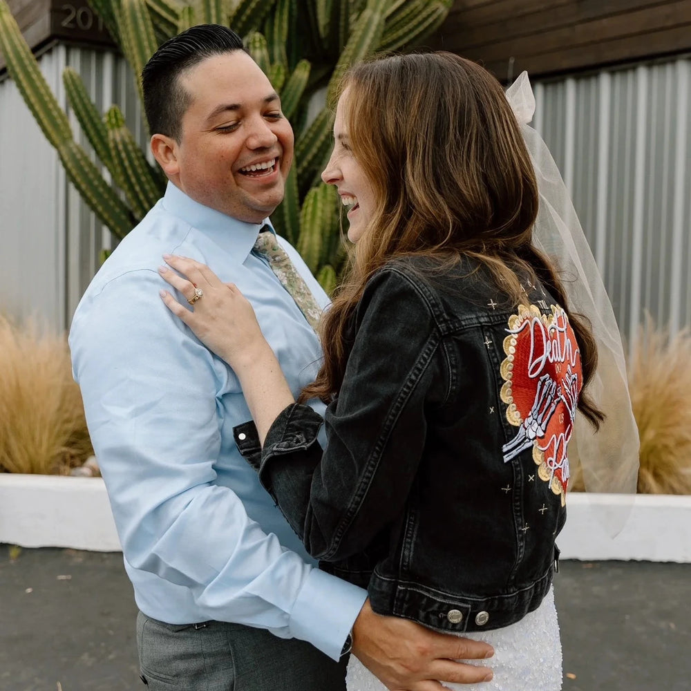 Bride and groom, laughing outdoors with a cactus behind them. The bride wears a black denim jacket with a "till death do us part" heart celestial desig including a skeleton pinky promise. 