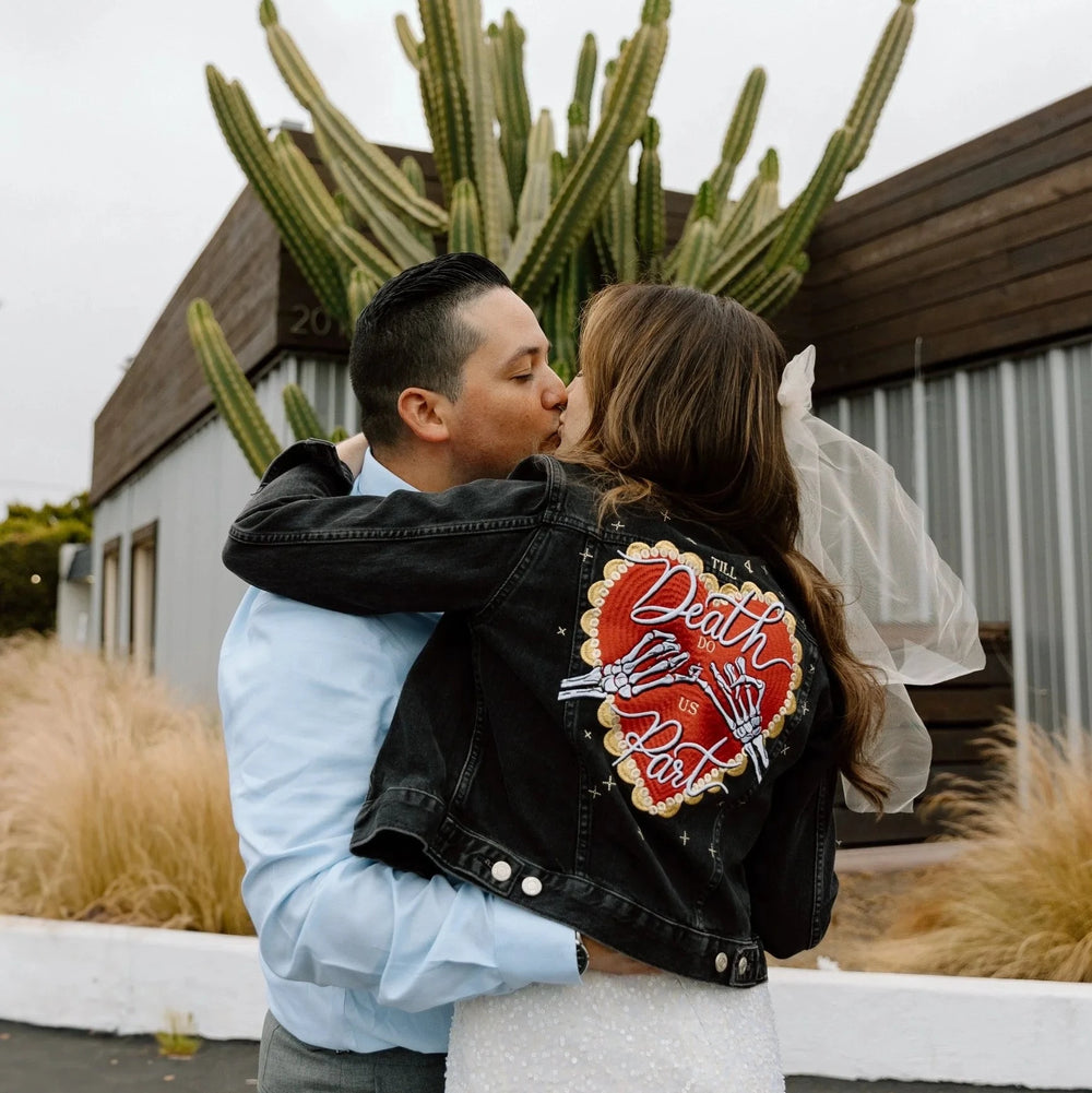 Couple just married, embracing outdoors in a cactus setting. The bride is wearing a white dress with a black denim jacket that has a till death do us part heart celestial design on the back. 