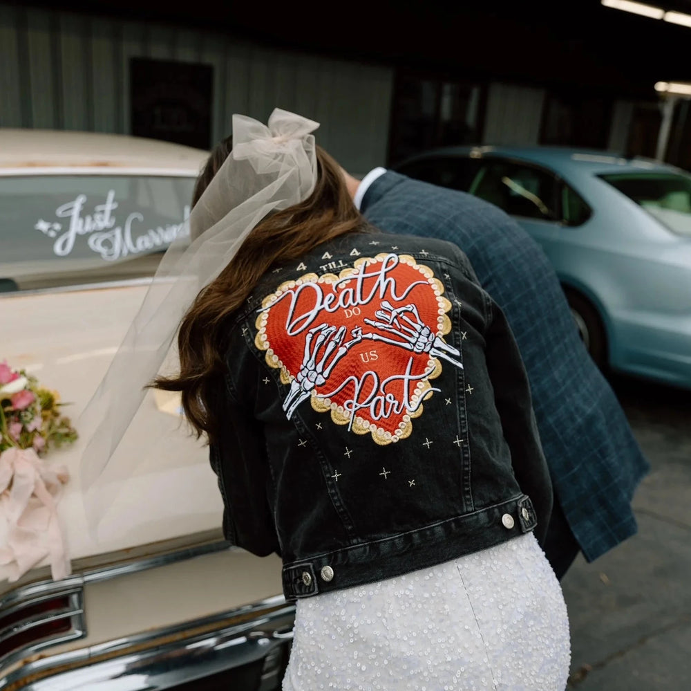 Bride wearing a bridal denim jacket with a bold heart "Till death do us part" design, standing next to a vintage car in a parking lot. She wears a bow veil and white wedding dress.