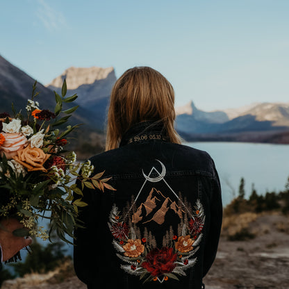 Close up of bride wearing a custom embroidered diamond mountain floral black denim jean jacket against a glacier background and her fall florals are held next to the design.