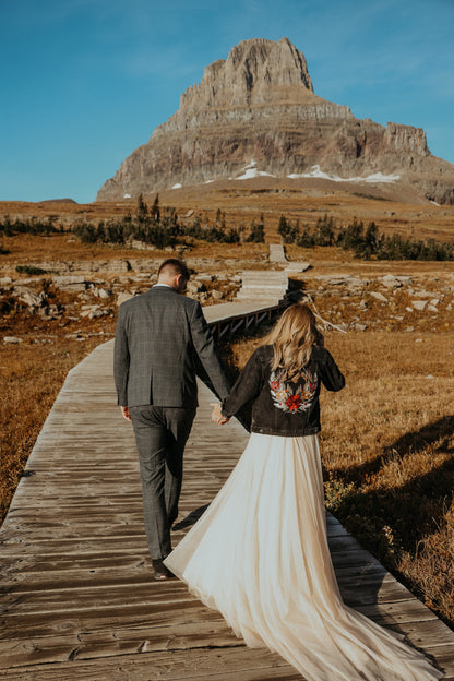 Couple holding hands on a wooden path with a mountainous landscape in the background. The bride wears a custom embroidered mountain floral design by Beth Wilson Embroidery