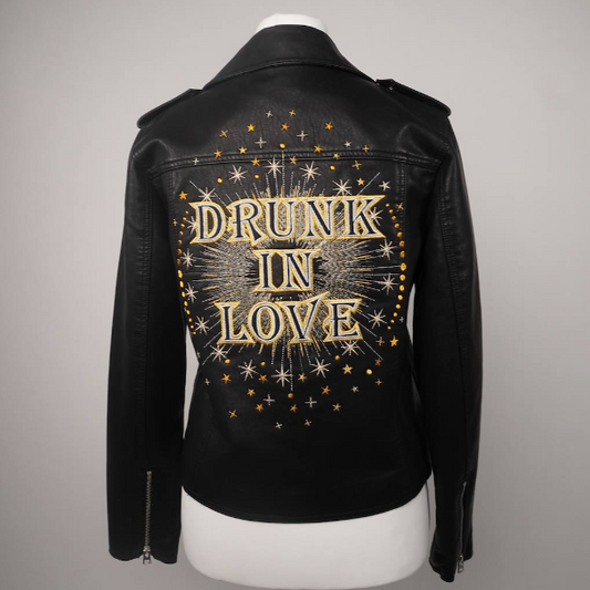 A black leather jacket displayed on a white mannequin torso, showing the back decorated with gold glittery text reading 'DRUNK IN LOVE' inside a circular starburst pattern.