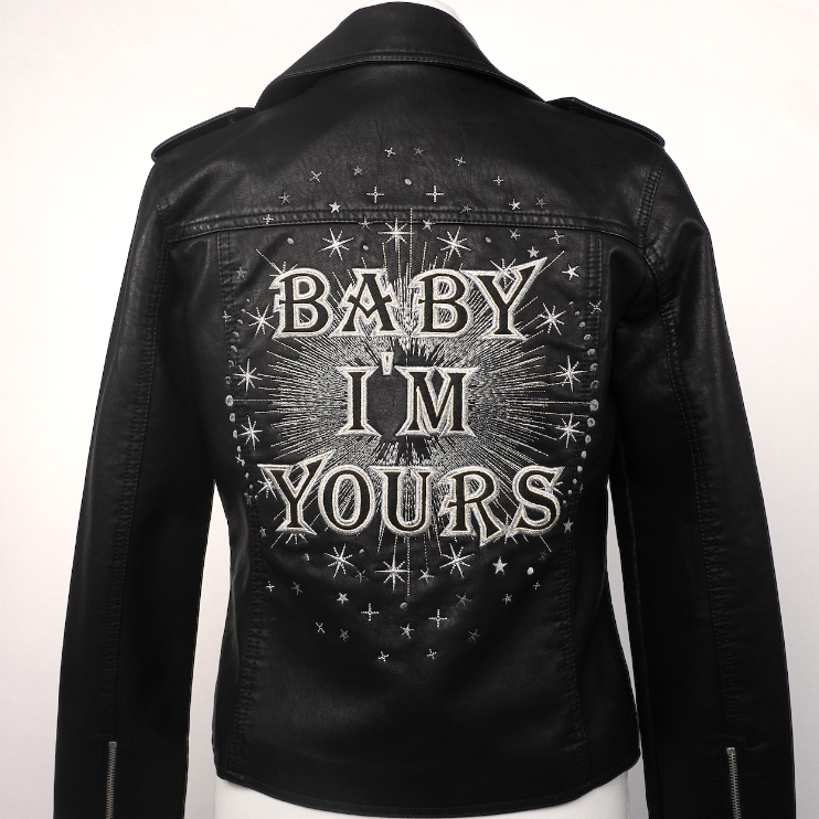 Black faux leather biker jacket with 'BABY I'M YOURS' and star graphics on the back, displayed on a white mannequin torso.