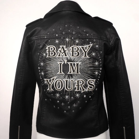 Black faux leather biker jacket with 'BABY I'M YOURS' and star graphics on the back, displayed on a white mannequin torso.