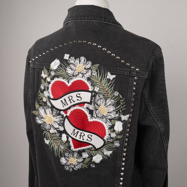 A black denim jacket displayed on a mannequin from the back, featuring a large embroidered design with two red hearts, white flowers, green leaves, and a banner reading 'MRS. MRS.