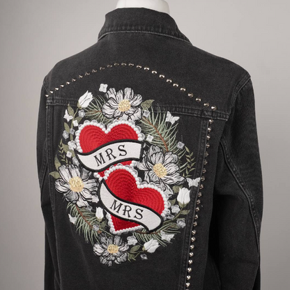 A black denim jacket displayed on a mannequin from the back, featuring a large embroidered design with two red hearts, white flowers, green leaves, and a banner reading 'MRS. MRS.