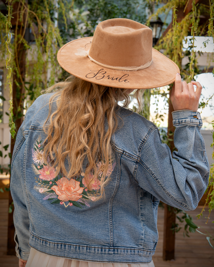 A woman viewed from behind wearing a wide-brim tan hat with 'Bride' embroidered on the brim, a light blue denim jacket with pink floral embroidery on the back, and a cream dress. She is posing in a garden setting with hanging greenery.