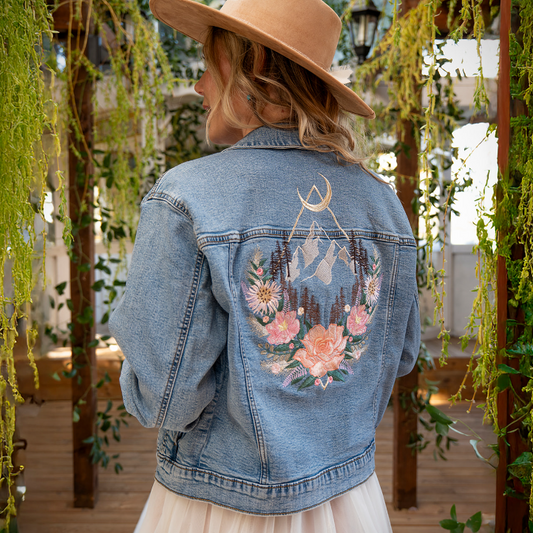 A woman viewed from behind wearing a light blue denim jacket with a large embroidered mountain and floral design on the back, a beige wide-brim hat, and a white tulle skirt, standing in a garden-themed setting with hanging greenery.