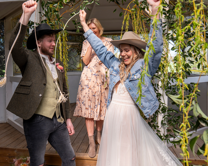 A bride in a white wedding dress and denim jacket, a groom in a brown blazer and cowboy hat, and a mother standing behind them on a porch, all holding a rope garland under hanging greenery.