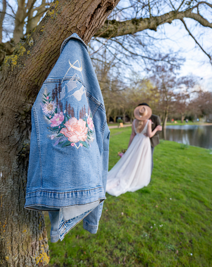 A light blue denim jacket with pink and white floral embroidery hangs from a tree trunk in a park, with a bride in a white wedding dress and a man standing in the blurred background near a lake.