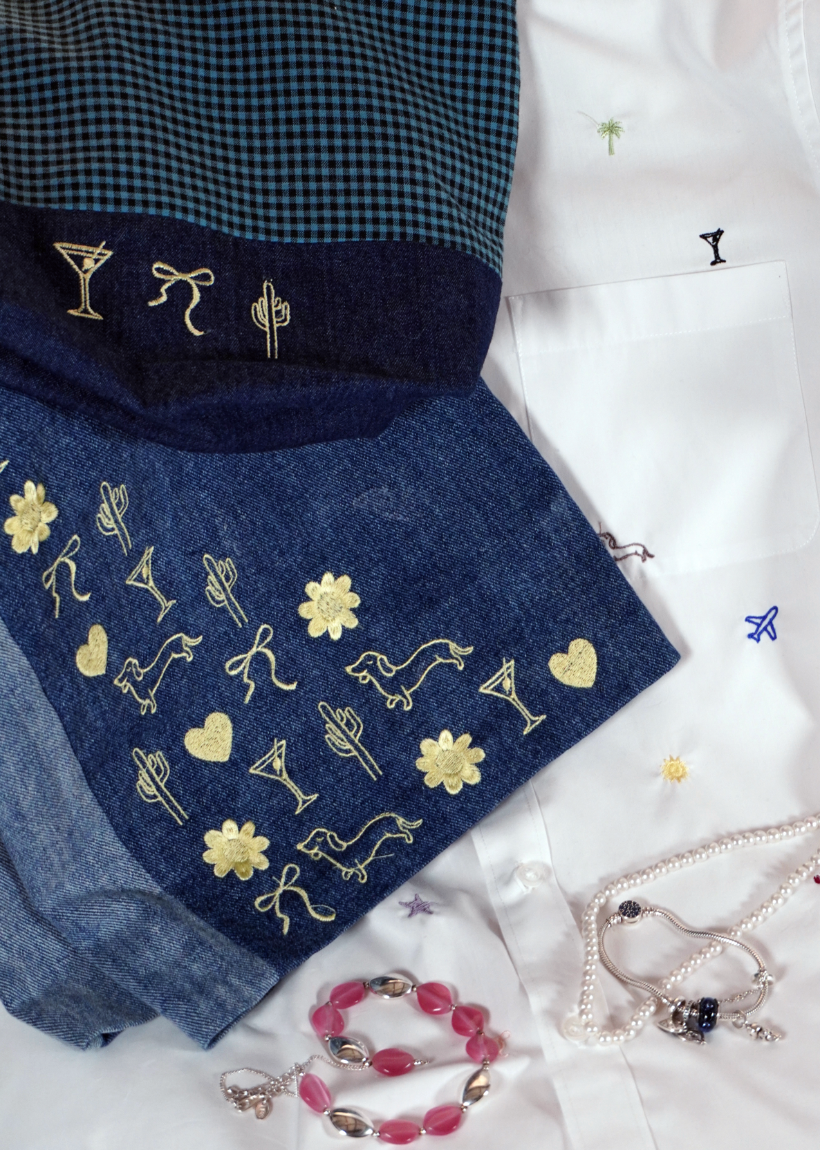 A styled flat-lay of a folded denim garment, likely shorts or a skirt, featuring a dark blue and black gingham pattern on the top section and a dark blue denim body with gold embroidered cocktail glasses, flowers, hearts, and abstract motifs. The garment is arranged on a white embroidered shirt with jewelry props like a beaded bracelet and a pearl necklace.