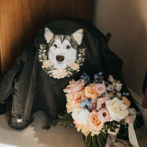A black denim jacket with a custom embroidered husky dog face and floral wreath design, styled next to a pastel flower bouquet on a beige floor against a wooden door.