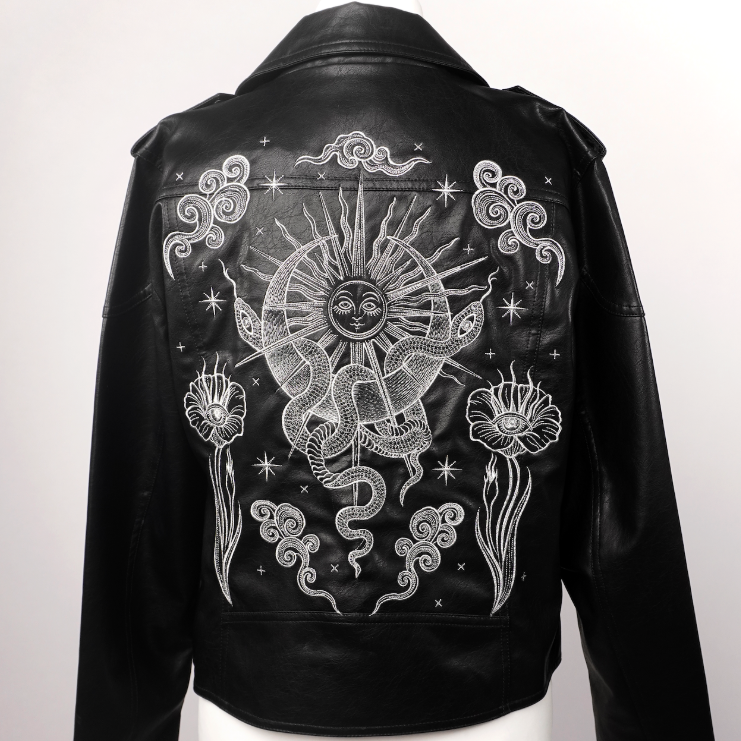 Black leather jacket displayed on a white mannequin torso, showing the back with a large white celestial graphic featuring a sun, snake, stars, and floral motifs.
