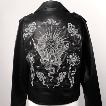 Black leather jacket displayed on a white mannequin torso, showing the back with a large white celestial graphic featuring a sun, snake, stars, and floral motifs.