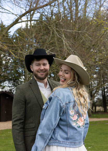 A couple outdoors in front of trees, with the woman wearing a light blue denim jacket featuring pink floral embroidery on the back, a beige cowboy hat, and a white dress. The man wears a black cowboy hat and a green blazer.