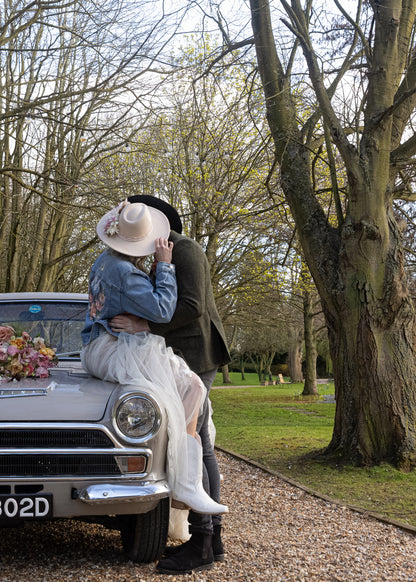 A couple embracing and kissing while sitting on the front of a vintage silver car on a gravel path, surrounded by trees in a park setting.