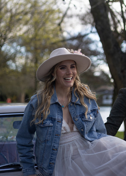A woman outdoors wearing a wide-brimmed light beige hat with a small flower accent, a blue denim jacket, and a white tulle dress, smiling while standing beside a car.