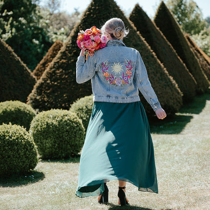 A woman stands outdoors in a garden wearing a light blue denim jacket with a colorful floral and sunburst embroidery on the back, paired with a long teal skirt. She is holding a vibrant bouquet of pink and orange flowers.