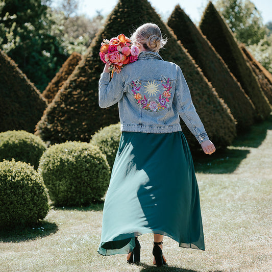 A woman stands outdoors in a garden wearing a light blue denim jacket with a colorful floral and sunburst embroidery on the back, paired with a long teal skirt. She is holding a vibrant bouquet of pink and orange flowers.