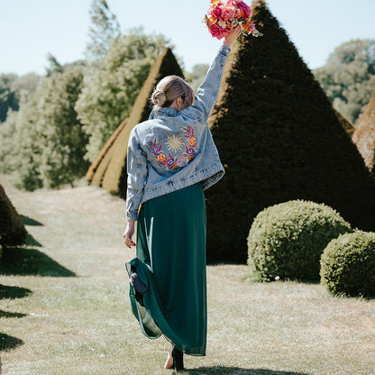 A woman stands outdoors in a garden wearing a light blue denim jacket with colorful floral embroidery on the back and a long dark green skirt. She is holding up a bouquet of pink and red flowers.