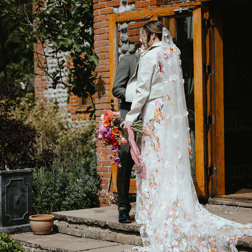A bride in a white floral lace wedding gown with a long train and veil stands on stone steps outside a brick building, holding a colorful bouquet next to a man in a gray suit.