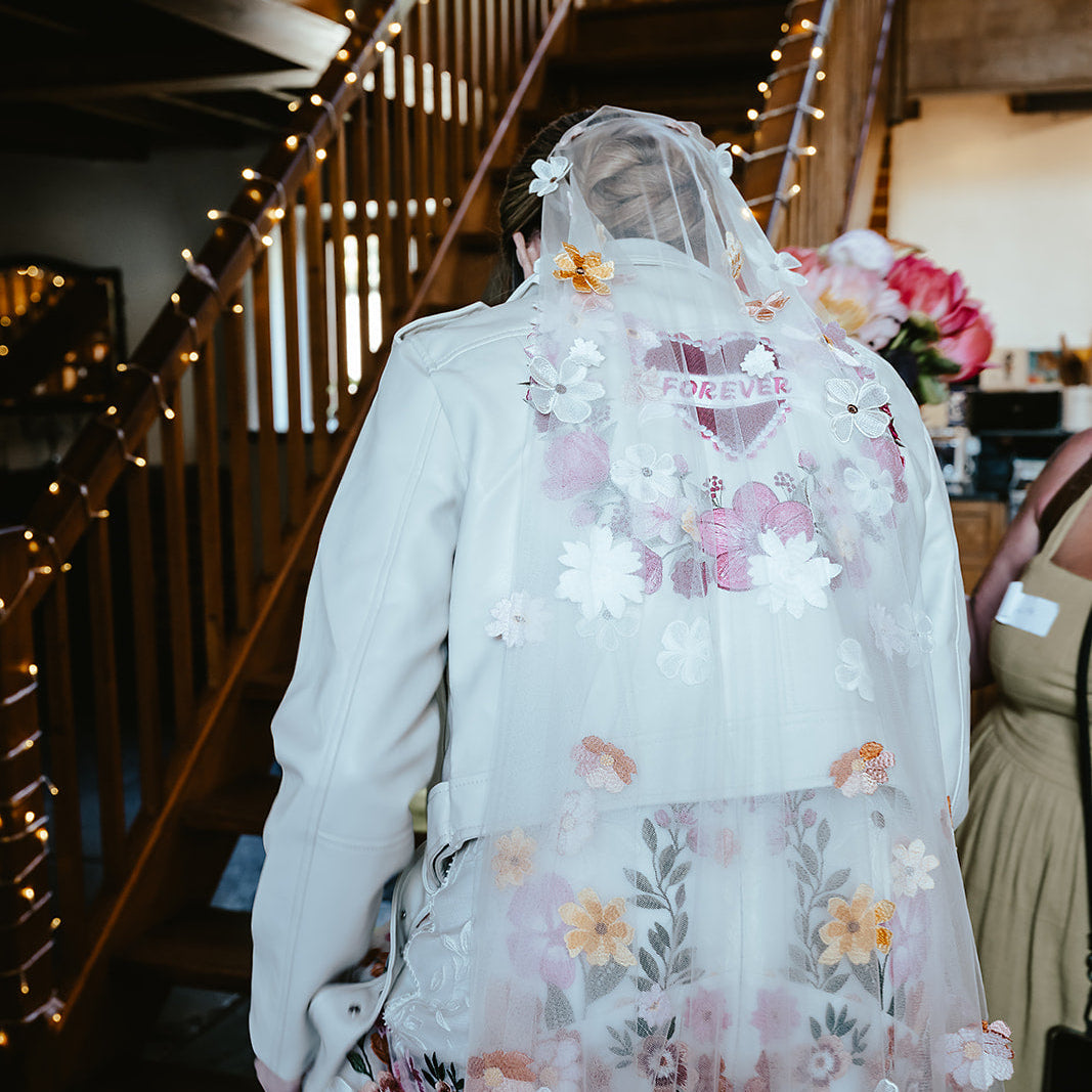A person viewed from behind wearing a long white bridal veil with large floral appliqués and a sheer tulle overlay decorated with pink, peach, and white flowers. The setting appears to be an indoor venue with wooden stairs and warm string lights in the background.