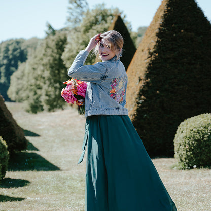 A woman stands outdoors in a garden wearing a light blue denim jacket with colorful floral embroidery on the back and a long teal skirt. She is holding a bouquet of pink flowers.