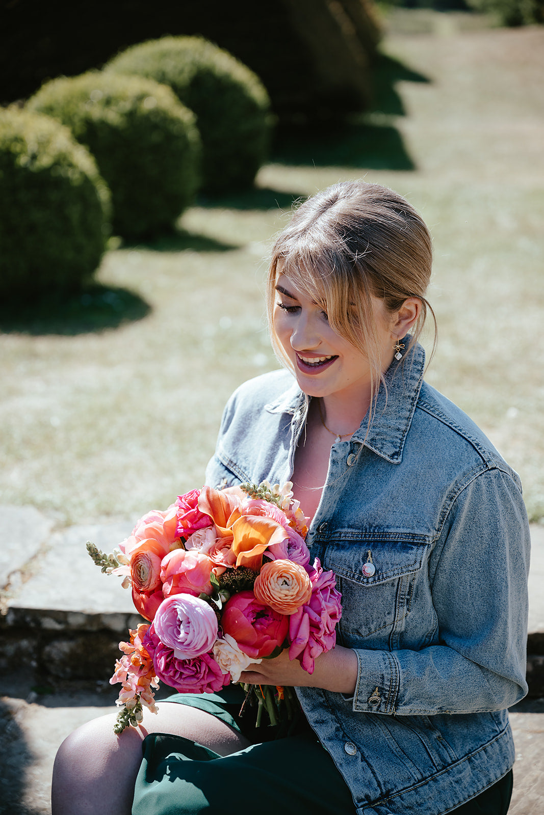 A woman sitting outdoors in a garden holding a vibrant bouquet of pink, orange, and purple flowers, wearing a light-wash denim jacket.