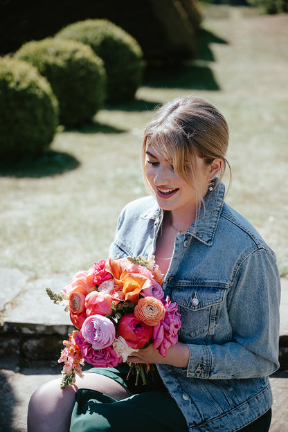 A woman sitting outdoors in a garden holding a vibrant bouquet of pink, orange, and purple flowers, wearing a light-wash denim jacket.