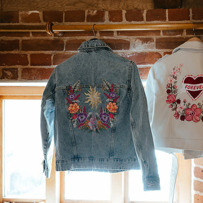 Two embroidered denim jackets hanging on a wooden rod in a rustic room with brick walls and a window. The left jacket is light blue with a colorful floral patch on the back, and the right jacket is white with a red heart and pink flowers on the back.