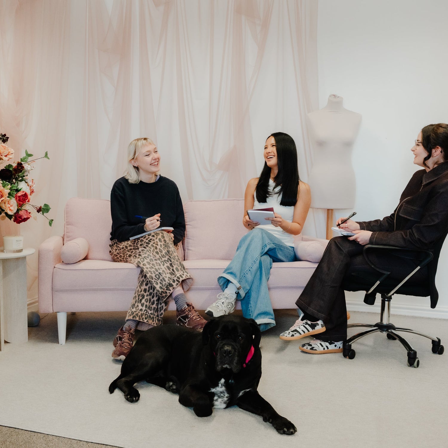 Three people sitting on a pink couch with a black dog in a room with a mannequin and flowers.